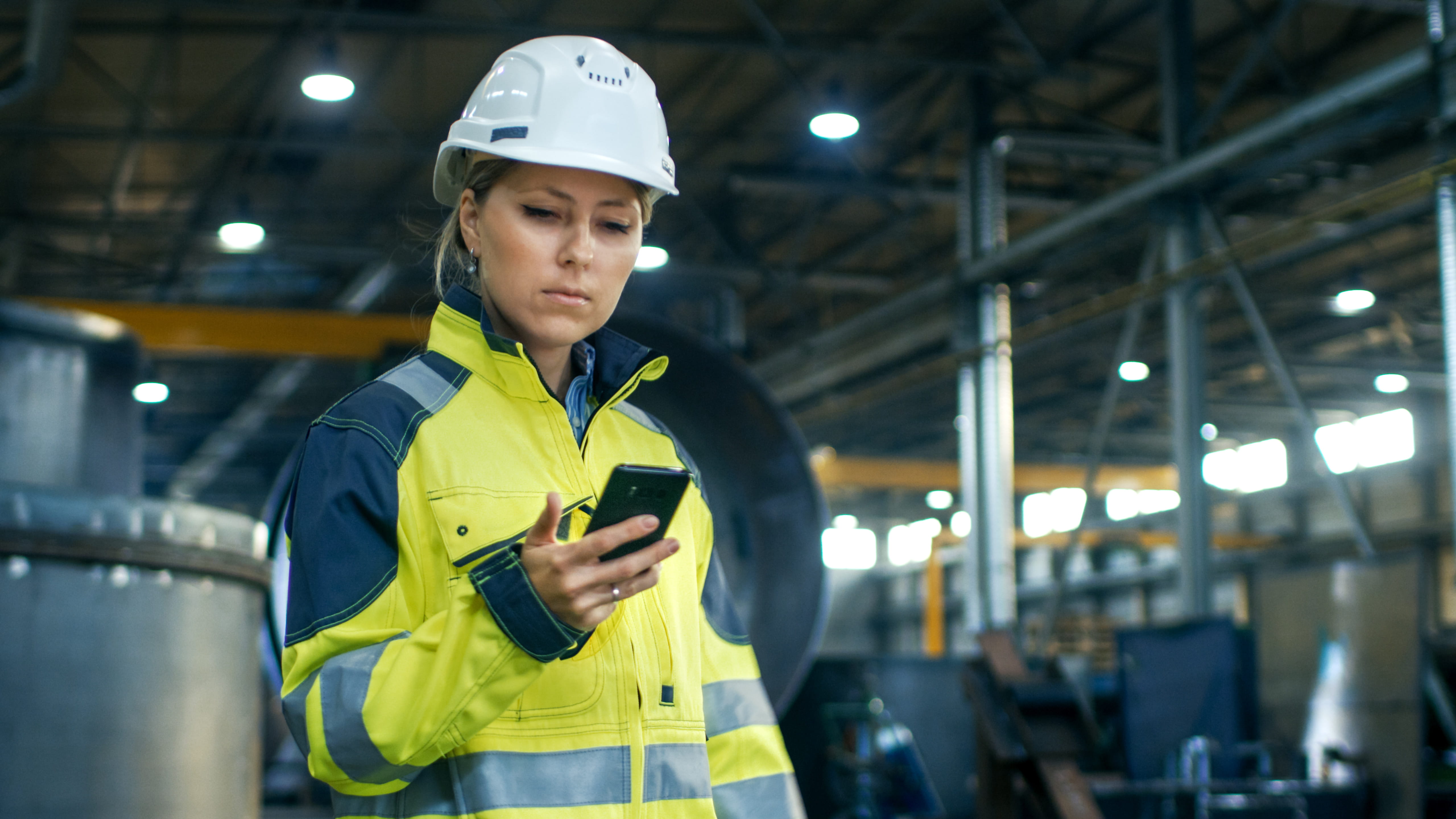 Female foreman in hard hat and safety vest checking her team's progress on her phone at a construction site.
